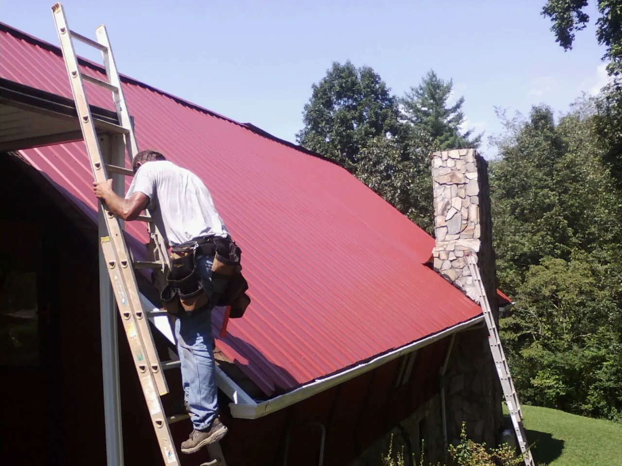 Roofer on a ladder beside a red metal roof and stone chimney — Soffit & Fascia Repair by Roofing Brothers in Escalon